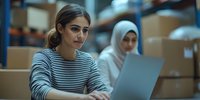 Young woman of South Asian descent wearing striped shirt working on laptop in warehouse with female colleague in hijab managing inventory and logistics operations.
