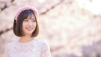 Young Asian woman with bob haircut wearing pink flower crown and white lace dress smiling during cherry blossom season in spring garden setting.