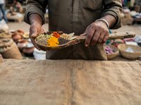 Vendor displaying colorful spice powder on wooden palette at outdoor market. Male hands holding turmeric and red chili spices for sale at traditional bazaar.