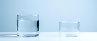 Two clear glasses on white surface against light blue background, one filled with water and one empty, representing hydration, consumption, and beverage concepts.