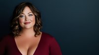 Confident plus size woman with curly brown hair wearing burgundy top smiling warmly against dark blue background for body positivity campaigns.
