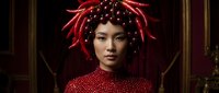 Young woman with pale complexion and East Asian features wearing elaborate red beaded headdress with dangling ornaments and matching red sequined garment against dark ornate background.