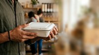 Man holding takeout food container in restaurant or kitchen setting, ready for delivery or meal service.