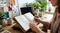 Young woman designer sketching in notebook at creative workspace with plants and coffee cup surrounded by inspiration materials and natural light.