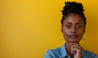 Young African American woman with natural hair in thoughtful pose against vibrant yellow background wearing denim jacket and gold earrings, contemplative expression.