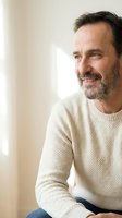 Middle-aged man with dark hair and beard wearing cream knit sweater, smiling while looking to side near bright window. Suitable for lifestyle, wellness, and portrait projects.