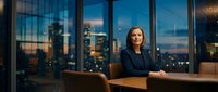 Professional middle-aged Caucasian businesswoman in dark suit sitting confidently at office desk with city skyline view through windows at dusk.