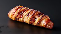 Fresh baked croissant drizzled with dark chocolate sauce and powdered sugar on dark background, close up view of flaky pastry with glossy glaze.
