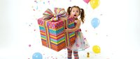 Young girl with light brown hair in pigtails, wearing colorful striped dress and socks, peeking from behind oversized rainbow gift box with confetti and balloons floating around on white background.