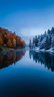 Serene lake reflecting autumn foliage and snow-covered evergreen trees under twilight sky, seasonal transition from fall to winter landscape.