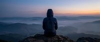 Hiker in dark jacket sitting on mountain peak at sunrise, overlooking misty valleys and distant hills during golden hour. Outdoor adventure and nature exploration.