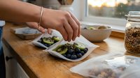 Person with medium skin tone preparing healthy breakfast with fresh berries and grains on wooden kitchen counter near window, suitable for wellness, nutrition and lifestyle content.