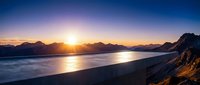 Dramatic sunrise over mountain range with golden sunlight reflecting on calm ocean waters and sandy beach in foreground, suitable for travel and nature backgrounds.