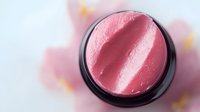 Pink shimmering powder highlighter or blush in round black compact container viewed from above on light background, showing luxurious cosmetic texture.