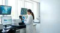 Female scientist in white lab coat analyzing data on multiple computer screens in modern laboratory with advanced technology and research equipment.