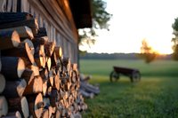 Stacked firewood pile against rural house wall at sunset, with wooden bench silhouette in blurred background meadow, creating cozy countryside atmosphere.