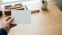 Hand holding blank white card at wooden desk with office supplies and stationery in background, suitable for branding and mockup projects.