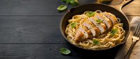 Sliced grilled chicken breast served over spaghetti pasta with fresh basil garnish in cast iron skillet on rustic wooden table for healthy dinner meal preparation.
