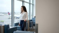 Young businesswoman with luggage waiting at airport terminal window overlooking runway with planes for business travel and departure lounge concepts.
