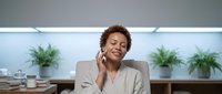 Woman with curly auburn hair, appearing to be in her 30s of mixed ethnicity, wearing a beige blazer, smiling while on a phone call in a modern office with plants and natural lighting.
