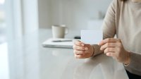Woman in beige sweater holding blank white card at desk with coffee cup, displaying mockup space for business message or promotional content.