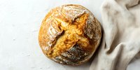 Freshly baked artisan sourdough bread with crispy crust and cross-cut pattern on white textured background with linen napkin.