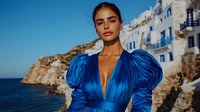 Young South Asian woman in luxurious blue satin dress with puff sleeves poses against Mediterranean coastal village backdrop with white buildings and ocean cliffs at sunset.