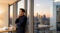 Thoughtful male professional in dark business suit standing by floor-to-ceiling windows in high-rise office, gazing at urban skyline at sunset, contemplating career decisions and business strategy.
