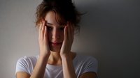 Young woman in white t-shirt covering face with hands showing signs of anxiety, depression, and emotional stress against dark background. Conceptual mental health photo.