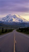 Straight asphalt highway with yellow center line leading toward snow-capped mountain peak at sunset, surrounded by evergreen forest and valley landscape for travel and adventure concepts.
