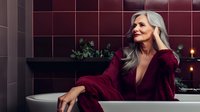 Elegant mature woman with silver gray hair relaxing in luxury bathroom wearing burgundy velvet robe. Wellness and self-care concept in dark moody interior setting.