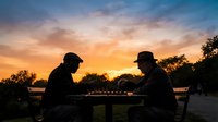 Two men in silhouette playing chess outdoors at sunset, with dramatic orange and blue sky, trees and nature background. Strategic game concept for leisure, competition, thinking.