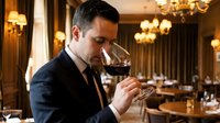 Professional sommelier in elegant suit tasting red wine in upscale restaurant with crystal chandeliers and refined dining atmosphere for hospitality industry.