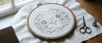 Delicate floral embroidery hoop with pink roses and green leaves on white fabric, surrounded by crafting supplies on linen cloth for needlework projects.