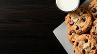 Fresh baked chocolate chip cookies on dark wooden background with glass of milk, overhead view of homemade dessert treats on napkin.