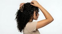 Woman with long curly black hair touching her head from side view, wearing beige top against white background for hair care and beauty.