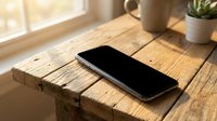 Modern smartphone with black screen lying on rustic wooden table near window with natural sunlight streaming in, minimalist workspace setup.