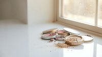 Broken makeup powder compacts scattered on white surface near window with natural light creating soft shadows and beauty product mess.