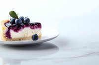 Slice of blueberry cheesecake with fresh berries on white plate against marble background. Creamy dessert topped with purple fruit sauce and mint leaf garnish.