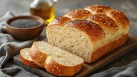 Freshly baked homemade bread with sesame seeds on wooden cutting board, sliced to show soft interior texture, served with honey and jam.