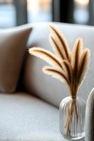 Dried pampas grass stems in ribbed glass vase against soft gray sofa background, creating minimalist home decor arrangement with natural beige tones.