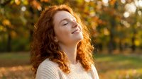 Young redhead woman with curly hair enjoying warm sunlight in autumn park with golden foliage, eyes closed in peaceful moment of relaxation and happiness.