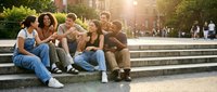 Diverse group of young adults sitting together on stone steps in urban park setting during golden hour sunset, sharing casual conversation and friendship moments.