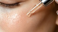 Closeup of serum dropper applying transparent skincare product to glowing, hydrated skin with visible pores and natural texture. Macro beauty shot.