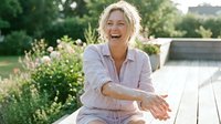 Mature woman with short blonde hair, Caucasian ethnicity, wearing lavender button-up shirt, laughing joyfully while sitting on wooden deck surrounded by blooming garden plants and green foliage.
