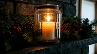 Glowing candle in a glass jar with LOVE cutout on a festive mantel with pine branches, pinecones, and red berries for holiday decor.