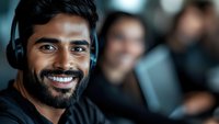 Male customer service representative with South Asian ethnicity wearing headset, smiling at camera in modern office environment with blurred colleagues in background.