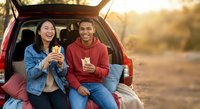Young diverse couple enjoying coffee break during road trip sitting in car trunk at golden hour sunset sharing drinks and laughing together outdoors.