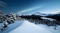 Snow covered mountain peaks and evergreen forest under starry night sky with moonlight illuminating pristine winter landscape in wilderness area.