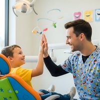 Young boy giving high five to male dentist in colorful scrubs during pediatric dental visit. Child wearing yellow shirt sits in dental chair smiling happily.